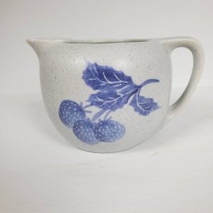 May include: A white ceramic creamer with a blue and white strawberry design. The creamer has a speckled glaze and a rounded body.