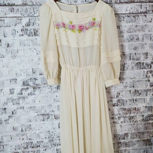 Op de afbeelding: Een vintage crèmekleurige jurk met een vierkante halslijn en lange mouwen. De bodice heeft roze en groene bloemenborduurwerk. De jurk heeft een geplooid taille en een zwierige rok, hangend aan een houten hanger.