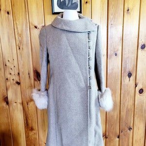 May include: A light gray vintage coat with a wide collar and button closure. The coat features long sleeves with faux fur cuffs. The coat is displayed on a mannequin against a wooden background.