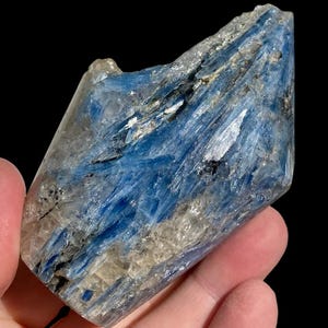 Pode incluir: Um espécime de cristal de cianita bruta apresentando uma mistura de formações minerais em azul vibrante e branco translúcido. A pedra tem um formato alongado e irregular com estrias visíveis e inclusões escuras na estrutura.