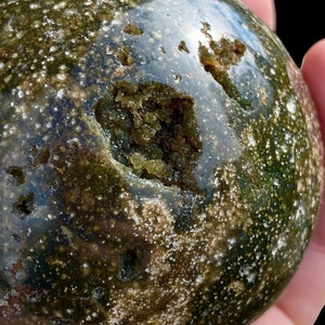 May include: A green and white sphere with a large, irregular opening revealing a cluster of smaller green crystals. The sphere has a smooth, polished surface and a speckled appearance.