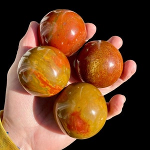 Può includere: Quattro sfere di diaspro rosso e giallo lucidate, ciascuna con un motivo unico di colori vorticosi. Le sfere sono lisce e lucide e sono tenute in una mano.