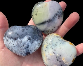 May include: Three heart-shaped stones in shades of gray, white, and yellow are held in a hand. The stones have a dendritic pattern, resembling tree branches. The background is black, highlighting the stones' colors and textures.