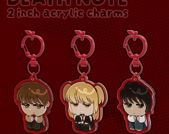 Może przedstawiać: Trzy 5-centymetrowe akrylowe zawieszki z postaciami w stylu chibi z Death Note. Każda zawieszka ma czerwone zapięcie w kształcie jabłka i czerwoną obwódkę. Postacie mają odpowiednio brązowe, blond i czarne włosy i siedzą w skulonej pozycji.
