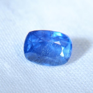 May include: A faceted, cushion-cut sapphire gemstone. The sapphire is a vibrant blue color and rests on a white fabric background. The gemstone's facets reflect light, creating a sparkling effect. This image showcases a precious stone.