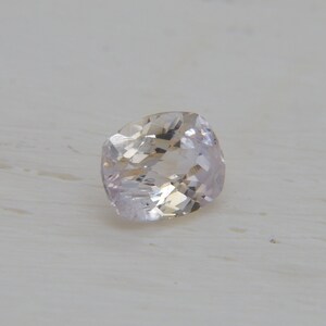 Puede incluir: Una gema facetada de color melocotón claro con talla cojín. La gema tiene un aspecto suave y translúcido y está colocada sobre una superficie blanca y texturizada. Las facetas de la piedra captan la luz, creando un brillo sutil.