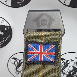 May include: Close-up of a tactical belt buckle with a Union Jack patch. The buckle is dark gray with a skull emblem. The belt strap is olive green with a woven texture. The patch features the British flag.