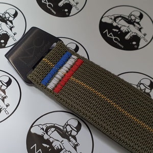 May include: Close-up of an olive green tactical watch strap with a black metal buckle. The strap features embroidered blue, white, and red stripes, and the buckle is engraved with "NXC Naguir de Combat".