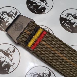 May include: An olive green nylon belt with a gunmetal buckle. The belt features embroidered stripes in black, yellow, and red. The buckle has an engraved design. The brand name "NXC STRAPS" is visible on the background.