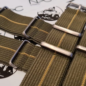 May include: Close-up of several olive green watch straps with a thin yellow stripe. Each strap has a silver-colored buckle. The straps are arranged on a white surface with a logo that says "NDC STRAPS".