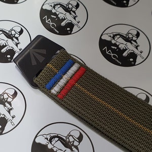 May include: An olive green nylon strap with a black buckle. The strap features embroidered stripes in blue, white, and red. The buckle has a geometric design. The background has repeating logos with the text "NAC STRAPS".