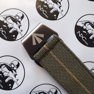 May include: Close-up of a tactical belt with a metal buckle. The belt is olive green with a thin gold stripe down the center. The buckle is silver with a triangular design. The background features multiple black and white logo stickers.