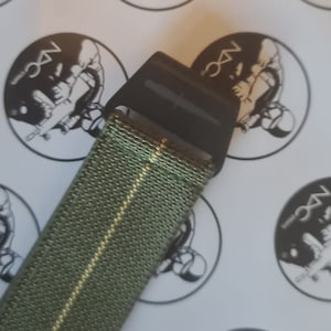 Puede incluir: Primer plano de un cinturón verde oliva con una hebilla negra. El cinturón tiene una textura tejida y una raya dorada en el centro. El fondo presenta pegatinas blancas con un gráfico en blanco y negro y el texto "NDC Straps".