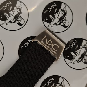 May include: Close-up of a black strap with a silver metal buckle. The buckle has the letters "NDC" and the words "Nageur de Combat" printed on it. The background features repeating black and white circular logos.