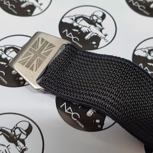 May include: Close-up of a black nylon strap with a silver-colored buckle. The buckle features an engraved Union Jack flag design. The strap is part of a watch band or similar accessory. The background includes circular logos with a soldier graphic and the text "NAC STRAPS".