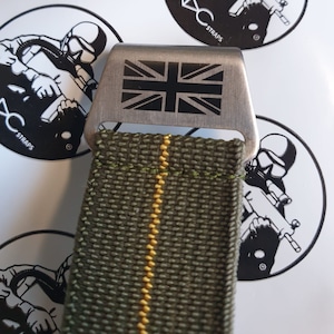 May include: Close-up of a green and yellow striped strap with a metal buckle. The buckle features the Union Jack flag. The strap is likely for a watch or similar accessory. The background includes a sticker with a graphic and the text "XC STRAPS".