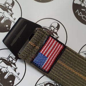 May include: Black metal belt buckle with a knife design, attached to an olive green belt with an American flag patch. The belt has a woven texture and a yellow stripe.