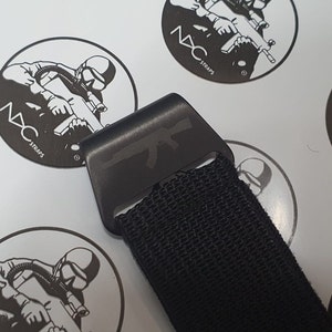 May include: Black strap with a black metal buckle featuring an etched AK-47 rifle design. The strap is made of a woven material. The background includes several white circular stickers with a soldier graphic and the text "NXC STRAPS".