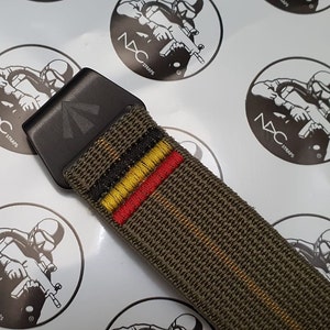 May include: Close-up of an olive green belt with a black buckle. The belt has embroidered stripes in black, yellow, and red. The buckle is black with a triangular design. The background features repeating stickers with the text "NXC STRAPS".