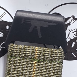 May include: A black metal buckle with a gun silhouette is attached to a green and tan woven strap. The buckle is rectangular with rounded edges. The strap has a repeating pattern of green and tan threads.