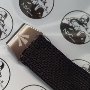 May include: Close-up of a black nylon belt with a silver-colored metal buckle. The buckle features a triangular design. The belt is set against a background with circular stickers that have a soldier graphic and the text "NXC STRAPS".