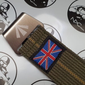 May include: A close-up of a watch strap with a metal buckle. The strap is olive green with a Union Jack patch. The buckle is silver with a white arrow design. The background has circular logos.