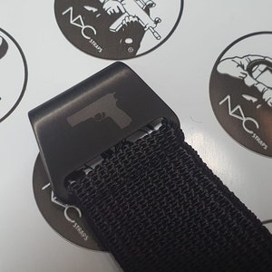 May include: Close-up of a black belt buckle with a gun silhouette. The buckle is attached to a black woven strap. The background features circular logos with the text "NXC STRAPS".