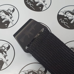 May include: Close-up of a black belt buckle with a gun design and a black nylon strap. The buckle has a matte finish. The background features a repeating pattern of a logo with a soldier and the text "NYC STRAPS".