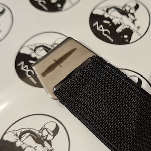 May include: A silver-colored metal buckle with a dagger design, attached to a black woven strap. The buckle is rectangular and features a detailed etching of a dagger. The strap is made of a tightly woven, black material. The background includes circular stickers with a soldier graphic.
