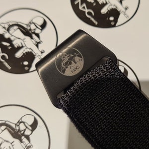May include: Close-up of a black metal belt buckle with a circular emblem featuring a soldier and a weapon, with the text "NXC" below. The buckle is attached to a black woven strap. The background includes additional circular logos.
