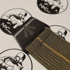 May include: Close-up of a black belt buckle with a sunburst design, attached to an olive green belt with a yellow stripe. The buckle is rectangular and has the text "NAC" on it. The belt is made of a woven material.