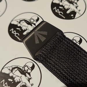 May include: Close-up of a black tactical belt buckle with a geometric design. The belt is made of a durable, woven material. The background features circular stickers with a soldier graphic and the text "NAC Straps".