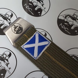May include: An olive green watch strap with a silver buckle featuring a soldier graphic and the text "NDC". The strap has a blue and white Scottish flag patch. The background has repeating "NDC STRAPS" logos.