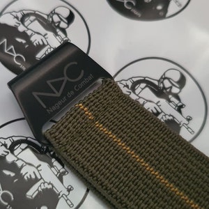 May include: Close-up of a watch strap with a dark green woven nylon band and a black metal buckle. The buckle is engraved with the letters "NAC" and the words "Nageur de Combat". The strap has a thin yellow stripe.