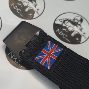 May include: Black nylon strap with a metal buckle. A small patch with the Union Jack flag is sewn onto the strap. The buckle has a skull design. The background has several white stickers with the text "NXC STRAPS".