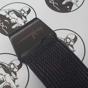 May include: Close-up of a black strap with a black buckle. The buckle has a gun silhouette etched on it. The strap is made of a woven material. The background has multiple white stickers with a black graphic and the text "DC STRAPS".