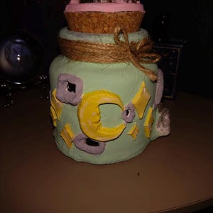 May include: A decorative jar with a light green base, adorned with yellow crescent moons and stars. The jar is topped with a pink layer, a cork base, and a small garden. A twine bow wraps around the jar.