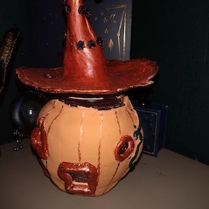 May include: A handmade, whimsical Halloween decoration. The piece features a pumpkin-shaped base with a witch's hat on top. The pumpkin is tan with brown accents and the hat is reddish-brown with black star embellishments.