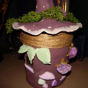 May include: A whimsical, handcrafted purple mushroom house with a pointed hat, white polka dots, and a scalloped brim. The house is adorned with green moss, faux flowers, and decorative leaves. A twine band encircles the middle.