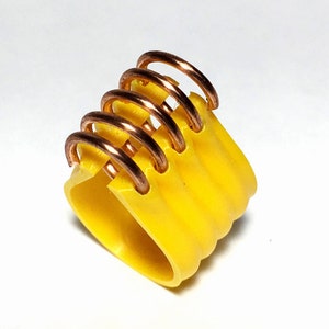 Puede incluir: Un anillo de plástico amarillo con una serie de anillos de alambre de cobre envueltos alrededor. Los anillos de cobre están apilados uno encima del otro y son visibles en la parte superior del anillo.