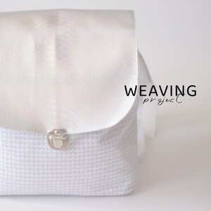 Puede incluir: Bolso tejido blanco con una solapa iridiscente plateada y un cierre plateado. El bolso tiene el texto "WEAVING project" impreso en la solapa.