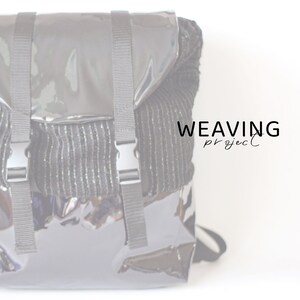 Può includere: Uno zaino nero con una finitura in pelle verniciata lucida e un pannello in tessuto intrecciato. Lo zaino ha spalline regolabili e una chiusura a fibbia. Il testo "WEAVING project" è stampato sul pannello in tessuto.