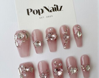 May include: Set of ten pink press-on nails with rhinestones and pearls. The nails are long and have a coffin shape. The nails are decorated with silver rhinestones and pearls. The nails are arranged in two rows of five. The nails are on a white background. The text "PopNailz EST. 2023" is visible in the top left corner of the image.