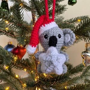 Picture of a crochet Christmas koala. The grey koala is holding a white Christmas star and is wearing a red Santa hat. The koala is facing forward and hangs on a decorated Christmas tree as an ornament