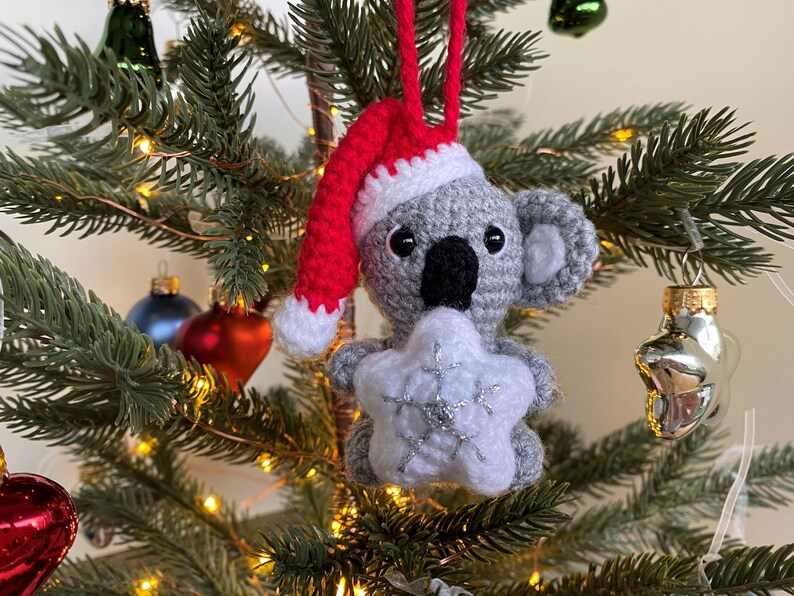 Picture of a crochet Christmas koala. The grey koala is holding a white Christmas star and is wearing a red Santa hat. The koala is facing forward and hangs on a decorated Christmas tree as an ornament
