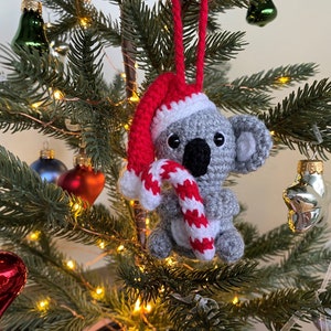 Picture of a crochet Christmas koala. The grey koala is holding a red and white candy cane and is wearing a red Santa hat. The koala is facing forward and hangs on a decorated Christmas tree as an ornament
