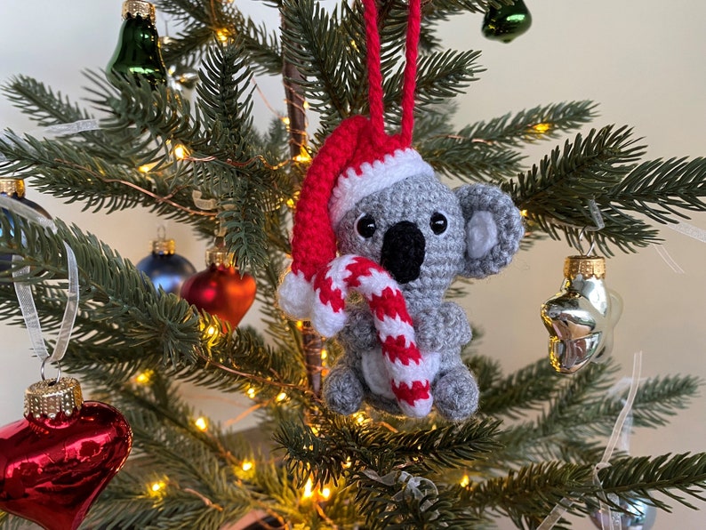 Picture of a crochet Christmas koala. The grey koala is holding a red and white candy cane and is wearing a red Santa hat. The koala is facing forward and hangs on a decorated Christmas tree as an ornament