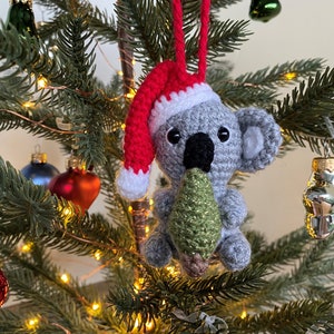 Picture of a crochet Christmas koala. The grey koala is holding a green Christmas tree and is wearing a red Santa hat. The koala is facing forward and hangs on a decorated Christmas tree as an ornament