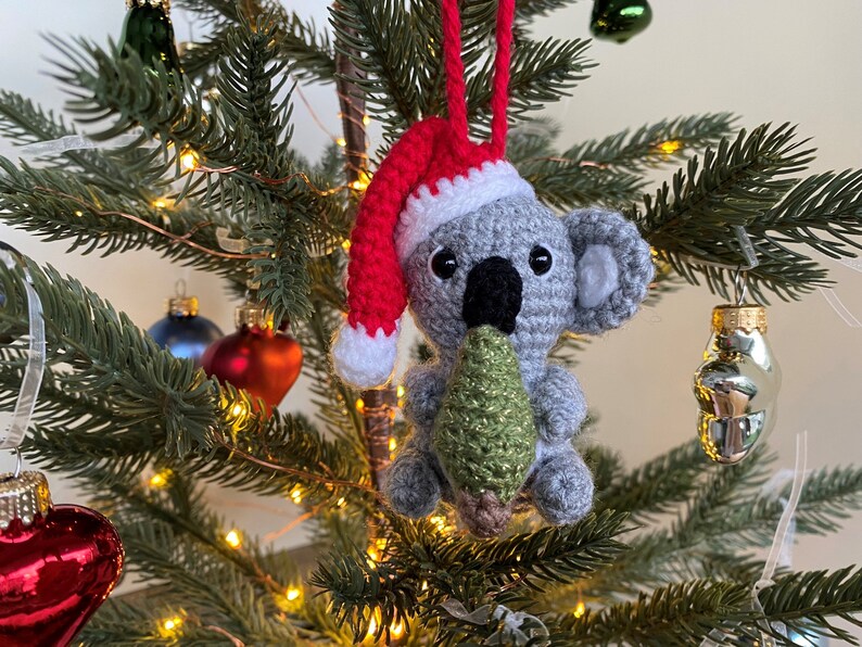 Picture of a crochet Christmas koala. The grey koala is holding a green Christmas tree and is wearing a red Santa hat. The koala is facing forward and hangs on a decorated Christmas tree as an ornament