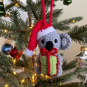 Picture of a crochet Christmas koala. The grey koala is holding a green and red christmas present and is wearing a red Santa hat. The koala is facing forward and hangs on a decorated Christmas tree as an ornament